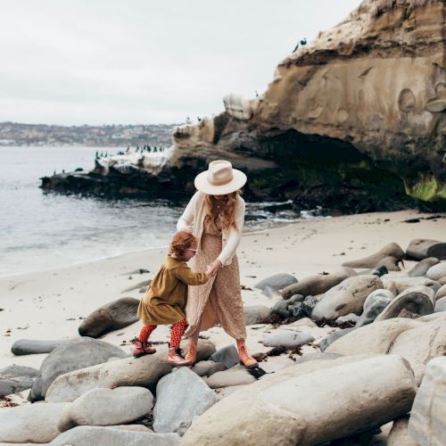 A woman in a hat holds a little girl on a rocky beach by the ocean beneath cliffs and waves.