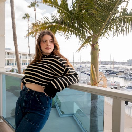 A woman in a striped sweater leans on a balcony railing, framed by palm fronds overlooking a marina of sailboats.