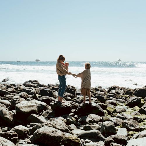 A family stands on a rocky shoreline, holding hands with a child as waves crash in the background, enjoying the seaside.
