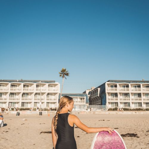 A woman in a black swimsuit stands on a sunny beach holding a pink surfboard, with hotel buildings and palm trees in the background.