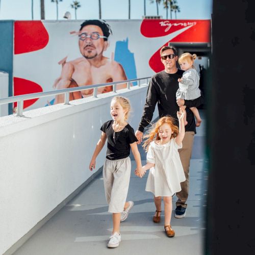 A family strolls down sunny stairs by a mural of a boxer, with two kids laughing and a man holding them close.