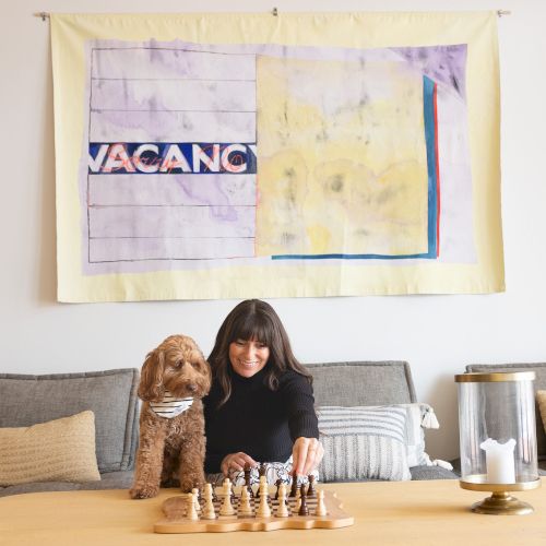A woman and a dog sit by a chessboard on a couch in a living room, with colorful wall art hanging above.