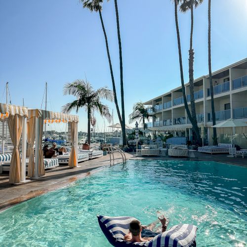 A sunny resort pool with palm trees and striped loungers, plus a man floating on an inflatable, with a marina in back.