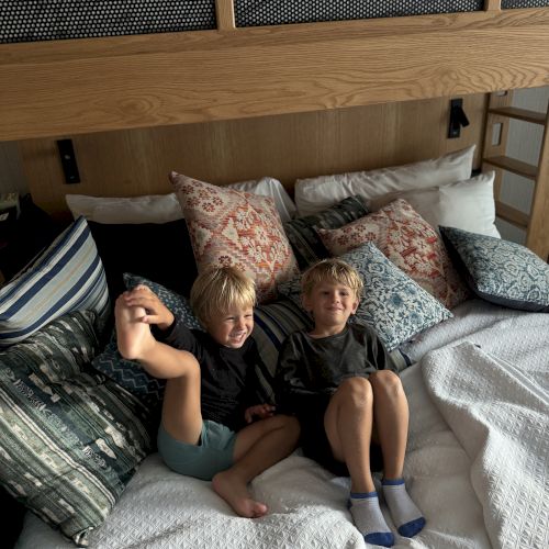 Two boys lie on a patterned bed with pillows and blankets, smiling at the camera in a cozy room with built-in paneling.