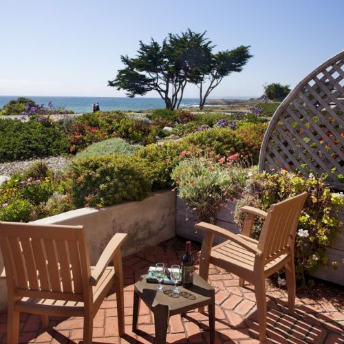 A sunny seaside garden patio with two chairs, a wine-and-glass table, blooming flowers, and an ocean view beyond trees.