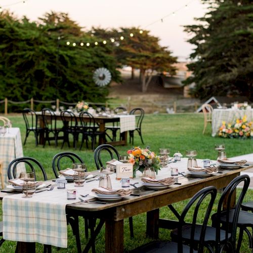 An outdoor garden dinner setup with long wooden tables, black chairs, place settings, flowers, and string lights overhead.
