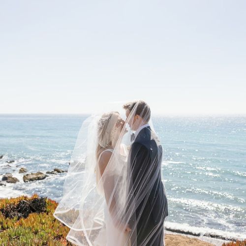 A bride in a flowing white veil stands on a sunny beach near the ocean, with sunlit waves sparkling in the background.