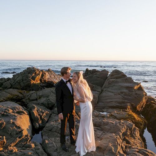 A couple in wedding attire stands on a rocky shore, gazing at each other as waves crash under a pastel sunset sky.
