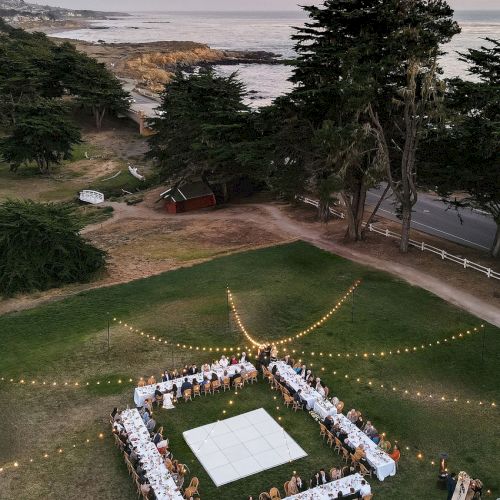 Outdoor seaside wedding dinner on a lawn with long tables, candles/string lights, and guests overlooking the ocean at dusk.