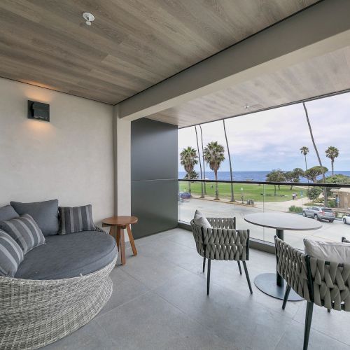 Modern patio living area with gray sofas and chairs, glass railing, and palm-lined ocean view outside.