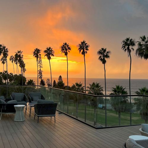 A sunset over the ocean with tall palm trees, viewed from a balcony patio with lounge chairs and a glass railing.