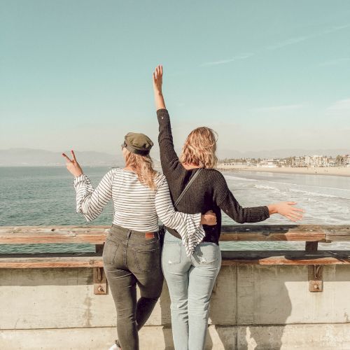 Two people on a seaside promenade with arms raised, enjoying the ocean view and a sunny coastal day.
