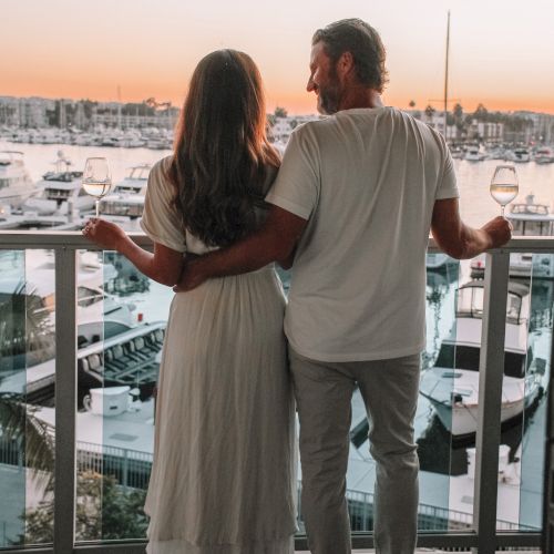 A couple on a waterfront balcony at sunset, holding wine glasses and embracing with a marina of boats behind them.