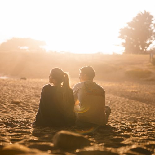A couple sits together on a sunlit beach at golden hour, facing the horizon as warm lens flares fill the scene.