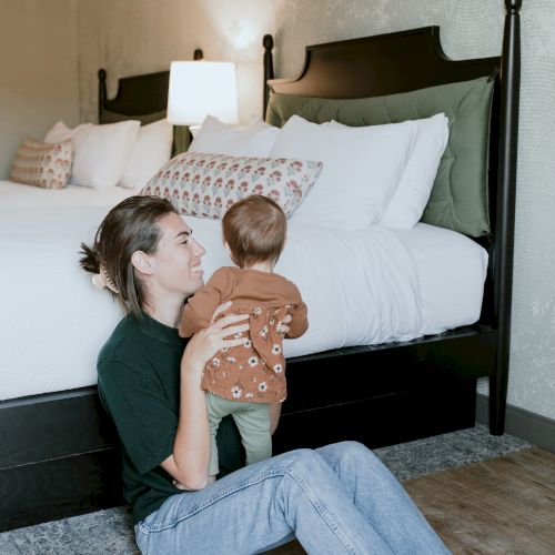 A parent cuddles a baby on the floor in a cozy bedroom while a bed with pillows sits behind them.