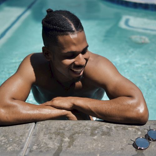A man smiles while lounging at a swimming pool, leaning on the edge as his sunglasses sit nearby.