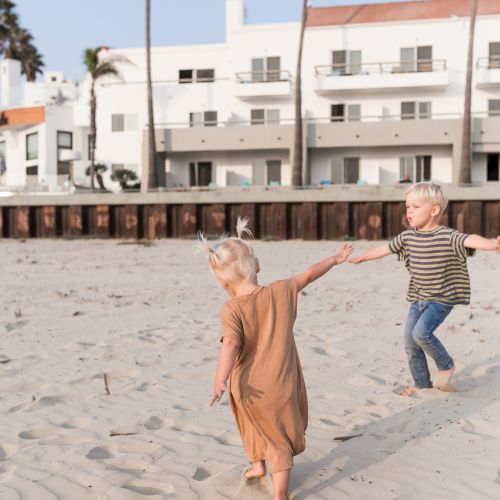 Two kids play on a sandy beach near palm trees and a white building with balconies, splashing and running in the sun.