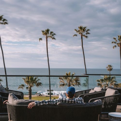 Oceanfront terrace with palm trees, a man relaxing on a couch, and waves under a cloudy sky.