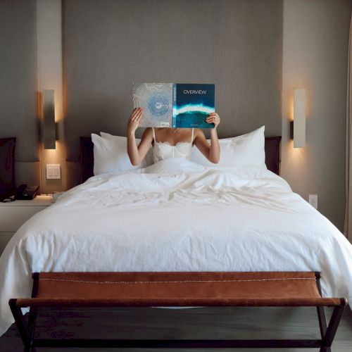 A person lounges in a hotel bed, covering their face with an &ldquo;OVERVIEW&rdquo; magazine, surrounded by white bedding and warm lamps.