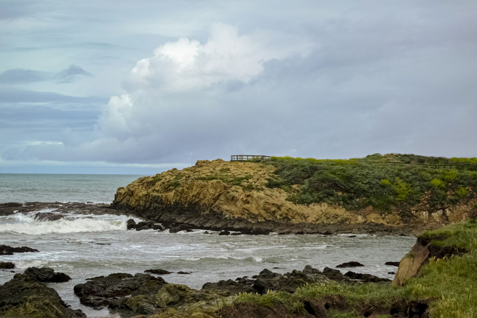 A cloudy ocean scene with churning waves crashing on rocky cliffs, lush green scrub, and a small building on the hilltop.
