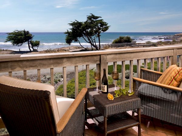 A seaside balcony with wicker chairs and wine glasses overlooking the blue ocean, trees, and rocky waves.