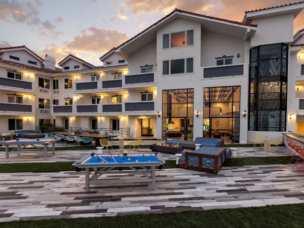 A luxury courtyard at dusk with a fire pit, billiards table, pool lounge chairs, and warm-lit modern apartments.