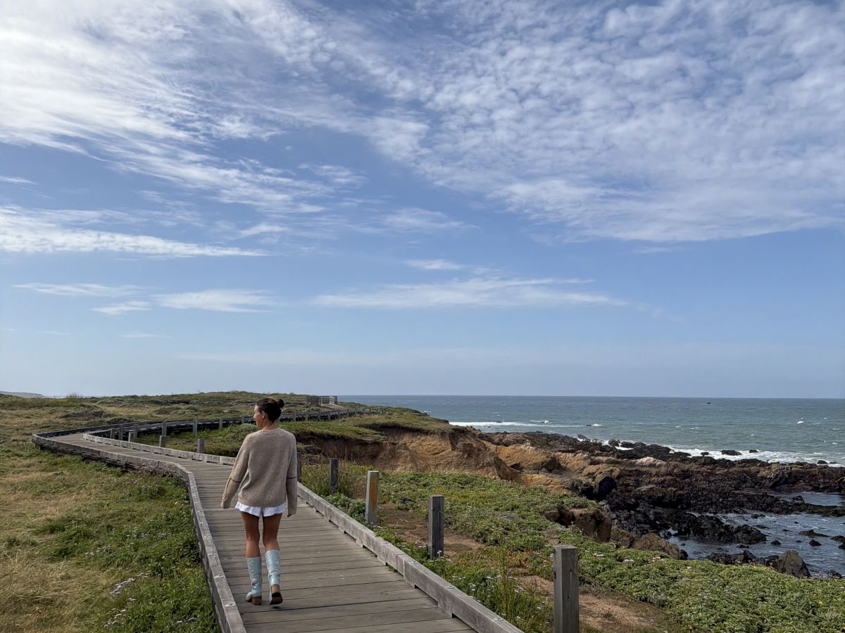 A person walks along a wooden boardwalk by the rugged ocean under a vast cloudy sky, soaking in the sea breeze.
