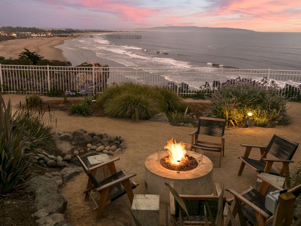 A beachfront sunset view with a bonfire, Adirondack chairs, and ocean waves rolling in under a colorful sky.