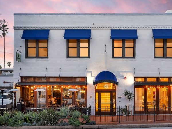 A white building with bright blue awnings and lit storefronts, palm trees, and a &ldquo;121&rdquo; entrance at sunset.