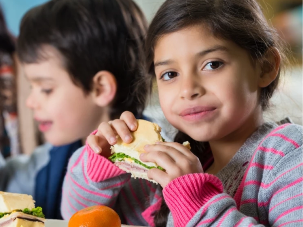 A girl in a striped sweater eats a sandwich while a boy sits beside her at a table.