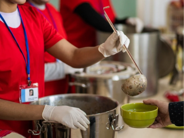 Volunteers in red shirts ladle food from large pots into a green bowl during meal service.