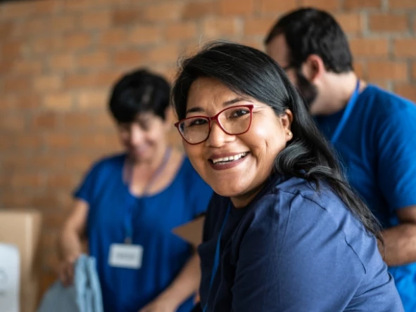 A smiling woman with red tilak and glasses chats at a community event with coworkers in blue shirts behind her.