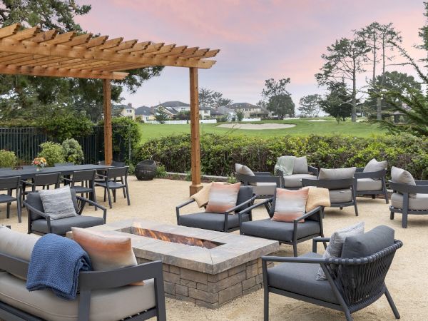 An outdoor patio with cushioned lounge chairs, a stone fire pit table, and views of a green lawn and trees at dusk.