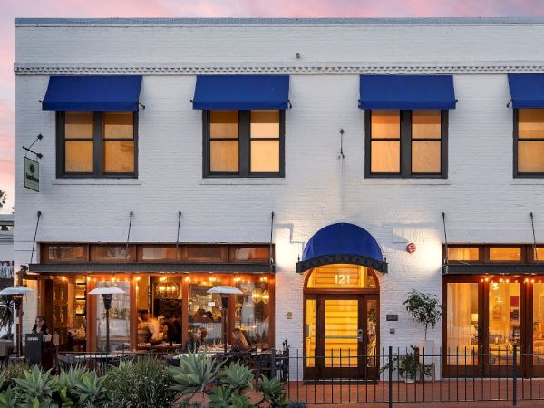 A white building with blue awnings and warm-lit storefronts at dusk, with palm trees framing the street.