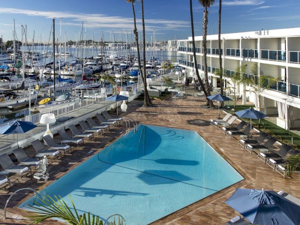 A seaside marina packed with sailboats beside a hotel pool, deck loungers, palm trees, and a modern multi-story building.
