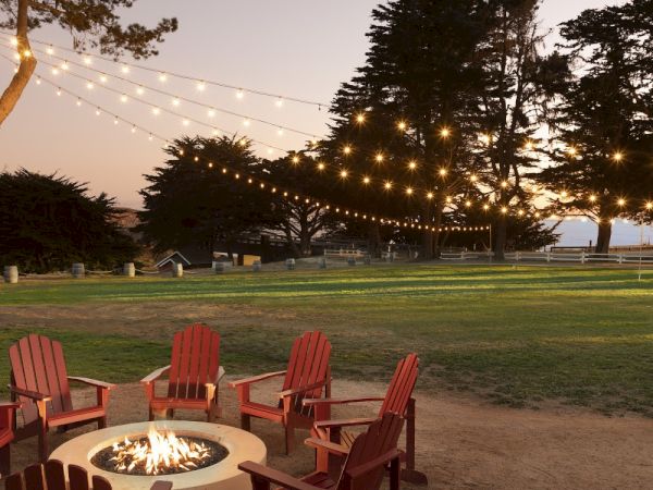 An outdoor firepit with red Adirondack chairs sits on a grassy lawn as warm string lights hang between trees at dusk.