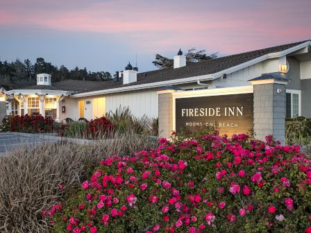 A seaside hotel exterior at dusk, with a &ldquo;Fireside Inn&rdquo; sign and bright pink flowers in front, under a colorful sky.