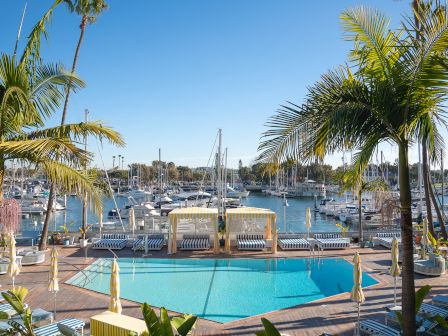 Sunny marina scene with a turquoise pool, striped loungers, and palm trees beside moored sailboats.