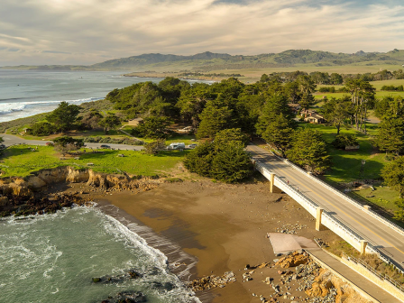 A coastal highway bridge spans a sandy beach by the ocean, with green hills, trees, and distant homes under warm sunset clouds.