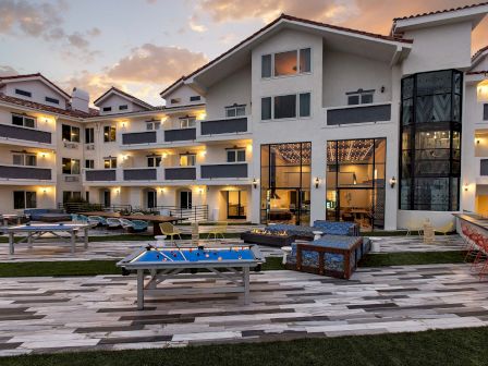 A luxury courtyard at dusk with a fire pit, billiards table, pool lounge chairs, and warm-lit modern apartments.