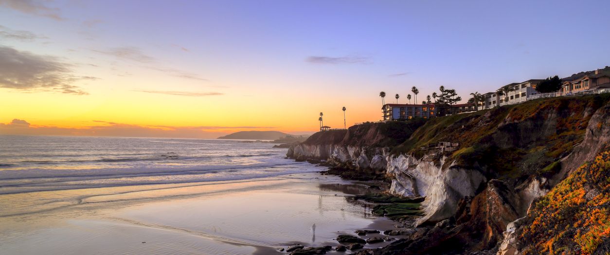 A sunset beach scene with gentle waves, rocky cliffs, palm trees, and hillside flowers beside homes overlooking the shore.