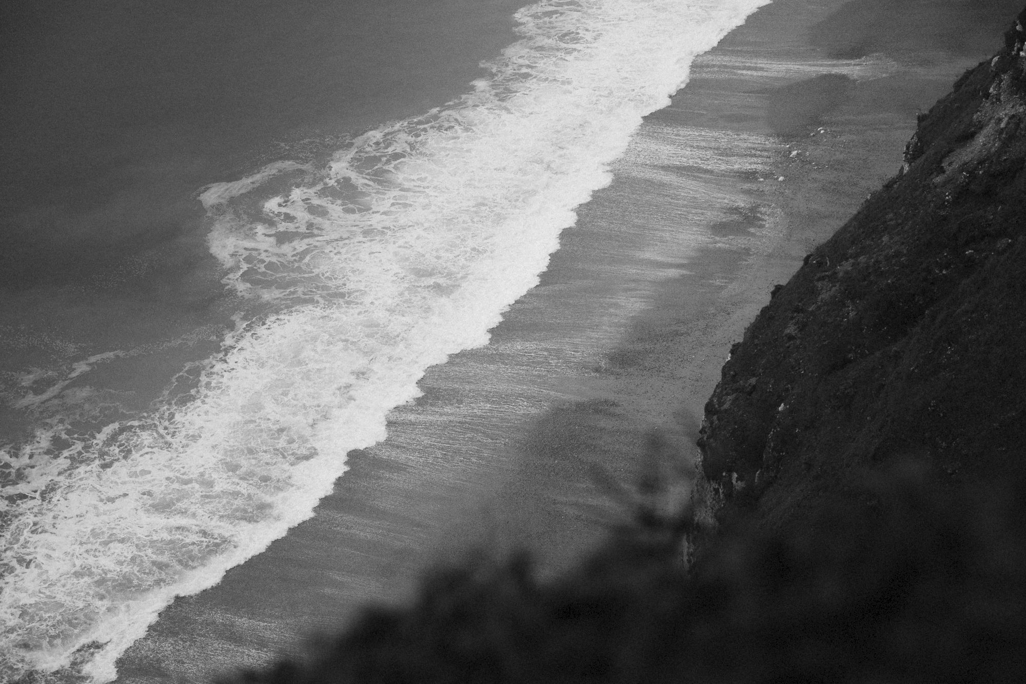 Black-and-white waves crash along a rugged cliff shoreline, foamy surf rolling into dark water.