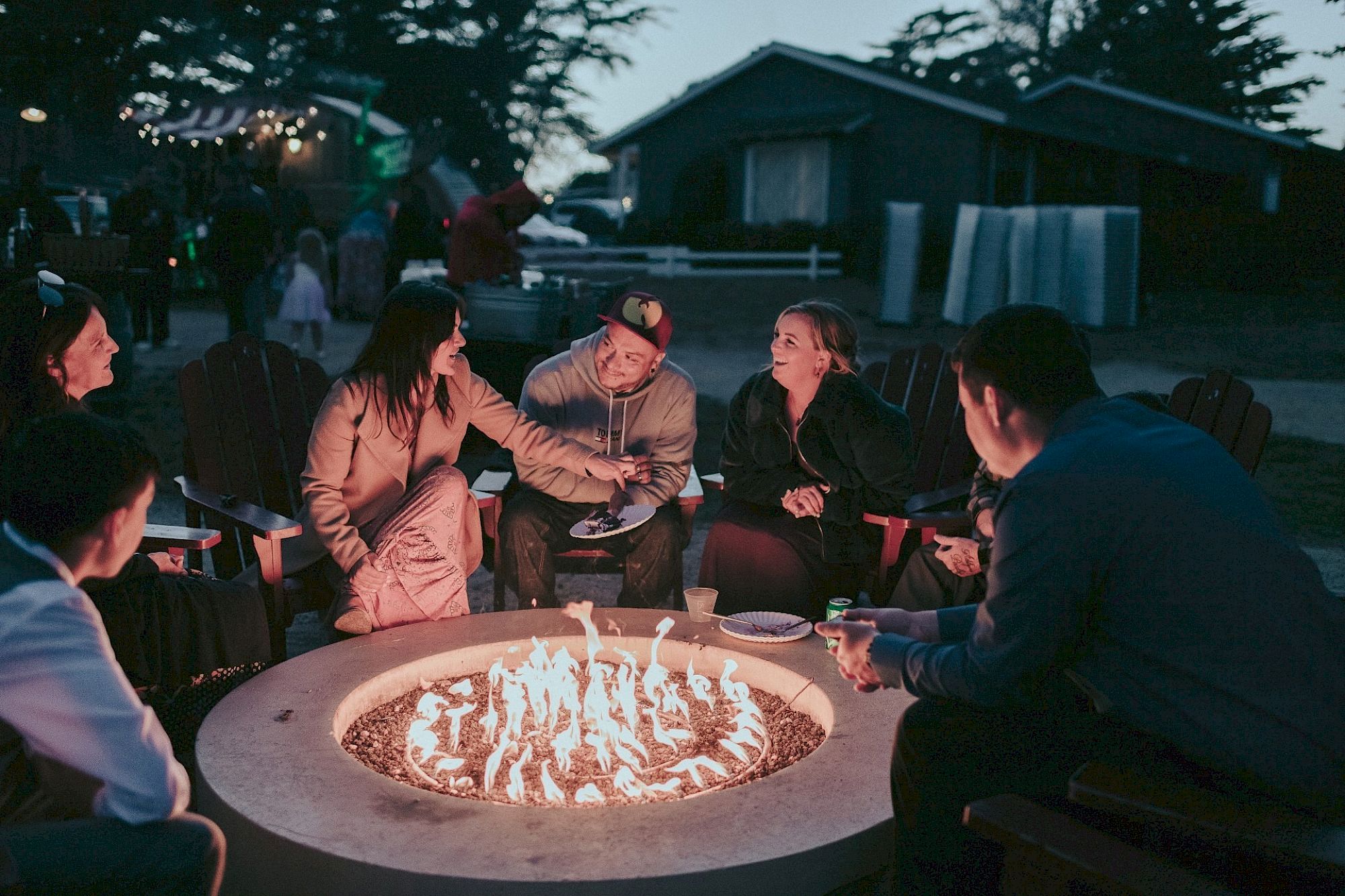 A group of people sit and chat around a backyard fire pit at dusk, roasting marshmallows and enjoying the warm flames.