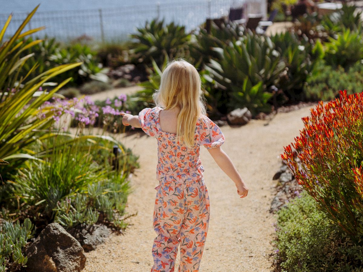 A little girl in a floral dress walks along a sandy path beside colorful coastal plants under a cloudy sky.