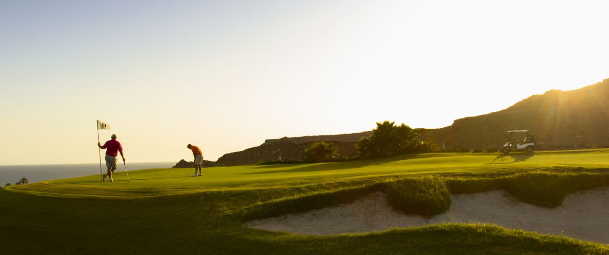 Golfers play on a sunny seaside course at golden hour, with dunes and a golf cart in the distance.