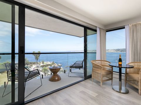 A modern seaside balcony/living room with wicker chairs, a small table with champagne, and floor-to-ceiling windows overlooking the ocean.
