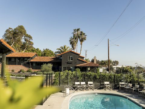 Sunny resort scene with a pool, patio chairs, palm trees, tiled roofs, and overhead power lines under a clear blue sky.