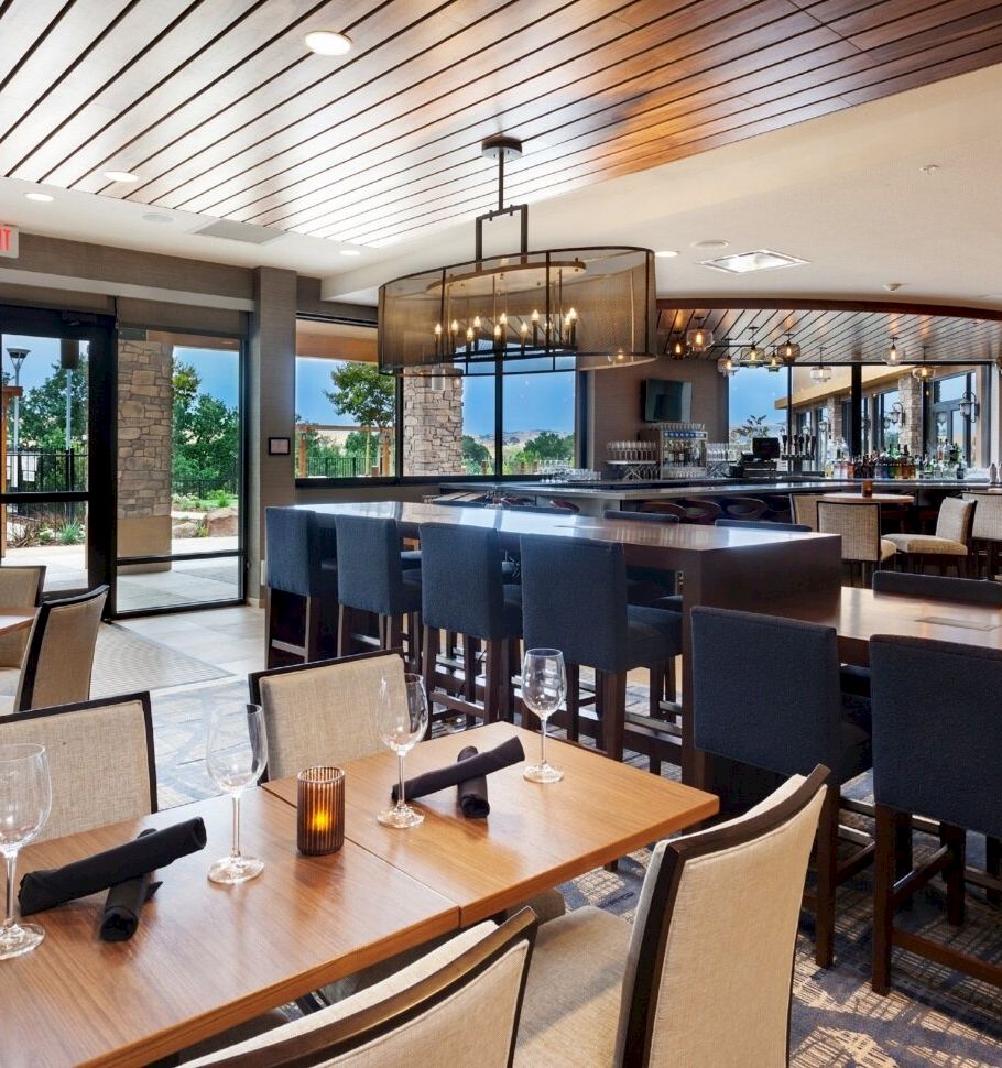 A modern restaurant interior with neatly set wooden dining tables, chairs, and a central bar under warm pendant lights.