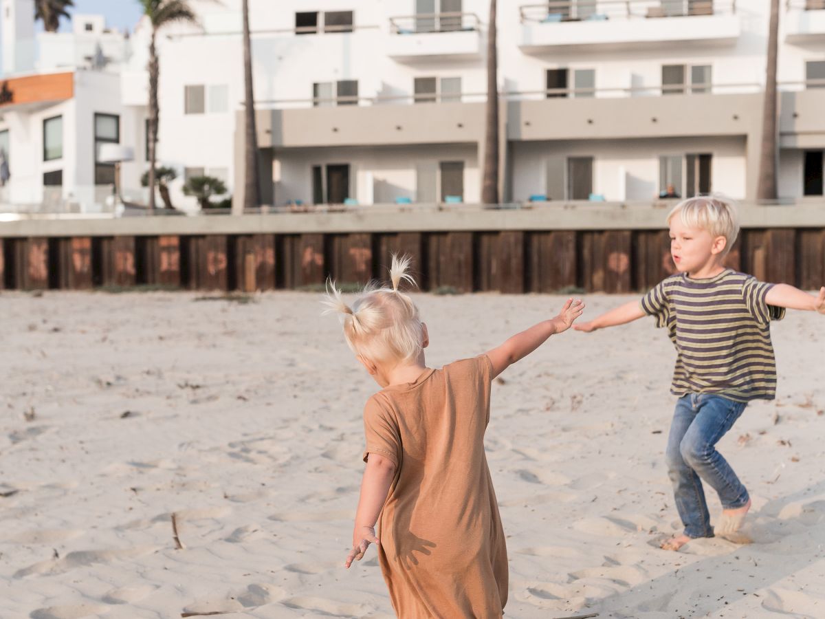 Two kids run and play on a sandy beach with palm trees and a white building in the background.