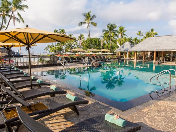 A tropical resort pool with sun loungers, umbrellas, and palm trees under a bright cloudy sky.